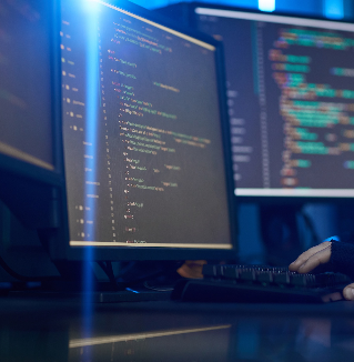 A person works on a desktop computer in a dark room, with two monitors displaying lines of code in a code editor.