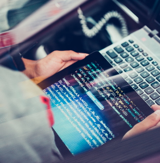 Close-up of someone holding a tablet displaying colorful programming code, with a laptop keyboard and some cables visible nearby.
